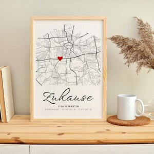 May include: A framed map print with a red heart marking a location. The print includes the word "Zuhause" in script, along with the names "Lisa & Martin" and coordinates. A white mug sits on a cork coaster.