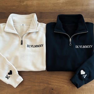 May include: Two matching sweatshirts, one cream and one black, with a quarter-zip collar. Each has the Roman numeral date IX.VI.MMXV embroidered on the chest. The cream sweatshirt has a 'K' and a heart on the sleeve, while the black one has an 'A' and a heart.