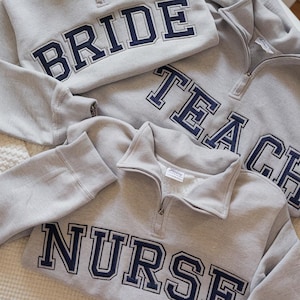 May include: Gray sweatshirts with navy blue lettering that reads "BRIDE", "TEACH", and "NURSE". Each sweatshirt has a quarter-zip collar. The sweatshirts are laying on a white surface.