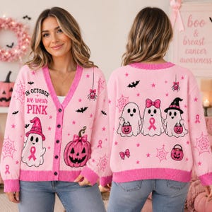 May include: A light pink V-neck button-down cardigan featuring a Halloween theme with ghosts, pumpkins, spiders, and pink breast cancer awareness ribbons. The front displays the text "IN OCTOBER we wear PINK" with a ghost wearing a pink hat.