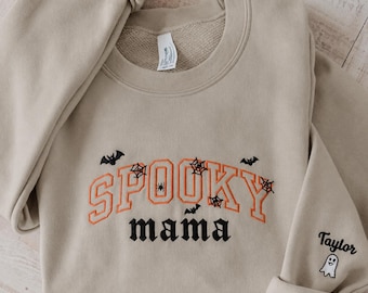 May include: A beige crewneck sweatshirt featuring orange embroidered text that reads SPOOKY and black embroidered text that reads mama. The design includes black spiderwebs, spiders, and bats. The sleeve features the name Taylor and a small ghost.