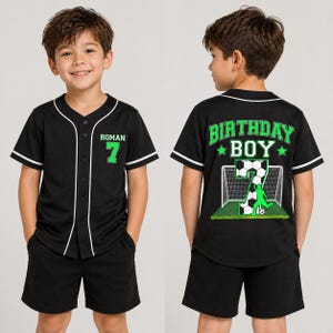 May include: Black baseball-style jersey and shorts set. The jersey has white trim, the name "ROMAN" and the number "7" in green. The back of the jersey says "BIRTHDAY BOY" with a soccer-themed graphic. The shorts are black.