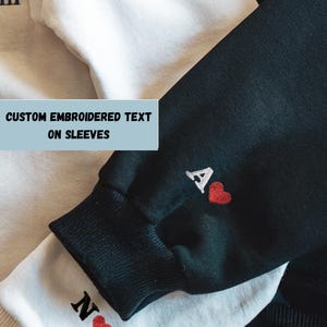 May include: Close-up of two sweatshirts, one white and one black, with embroidered details. The black sweatshirt cuff features a white "A" and a red heart. The white sweatshirt cuff has a black "N" and a red heart. Text on the image reads "CUSTOM EMBROIDERED TEXT ON SLEEVES."