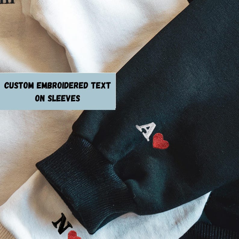 May include: Close-up of two sweatshirts, one white and one black, with embroidered details. The black sweatshirt cuff features a white "A" and a red heart. The white sweatshirt cuff has a black "N" and a red heart. Text on the image reads "CUSTOM EMBROIDERED TEXT ON SLEEVES."
