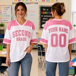 May include: A white and pink V-neck jersey with short sleeves and striped cuffs. The front features the pink text "SECOND GRADE" in a varsity font. The back displays the pink text "YOUR NAME" above the large pink number "00" in a varsity font.