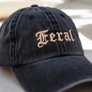 May include: Black baseball cap with the word "Feral" embroidered in a tan, gothic-style font. The cap has a curved brim and a distressed, washed appearance, suggesting a vintage or worn look. The cap is made of a soft fabric.