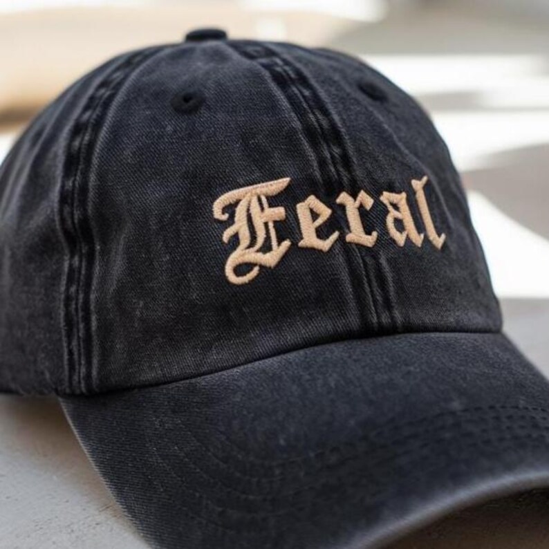 May include: Black baseball cap with the word "Feral" embroidered in a tan, gothic-style font. The cap has a curved brim and a distressed, washed appearance, suggesting a vintage or worn look. The cap is made of a soft fabric.