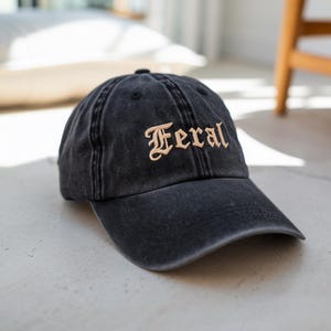 May include: A black, distressed baseball cap with the word "Feral" embroidered in a tan, gothic-style font across the front. The cap has a curved brim and a classic six-panel design. The hat is made of a soft, worn material.