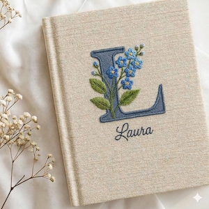 May include: A beige linen-textured journal with an embroidered blue floral design and the letter "L". The name "Laura" is embroidered below the letter. The journal is closed and resting on a white surface.