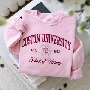 May include: A pale pink sweatshirt featuring the text "CUSTOM UNIVERSITY" in burgundy, arched across the chest. Below the arch is an emblem and the words "School of Nursing". The sweatshirt has a crew neck and long sleeves.
