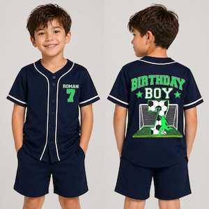 May include: Navy blue baseball-style shirt and shorts set. The shirt has white trim, the number 7 in green, and the name "ROMAN". The back of the shirt says "BIRTHDAY BOY" with a soccer-themed graphic. The shorts are navy blue.