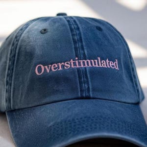 Op de afbeelding: Een blauwe denim baseballcap met het woord "Overstimulated" in roze geborduurd. De cap heeft een gebogen klep en een knoop aan de bovenkant. De hoed is een klassieke stijl, geschikt voor casual wear.