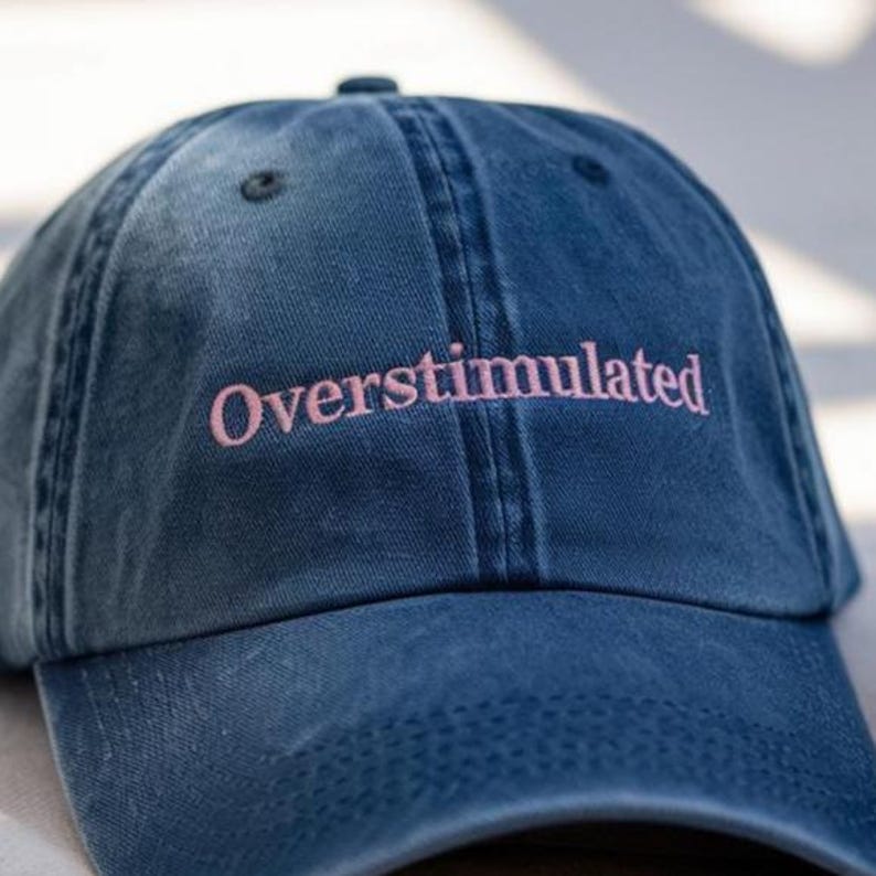Op de afbeelding: Een blauwe denim baseballcap met het woord "Overstimulated" in roze geborduurd. De cap heeft een gebogen klep en een knoop aan de bovenkant. De hoed is een klassieke stijl, geschikt voor casual wear.