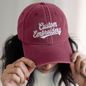 May include: A burgundy baseball cap with the words "Custom Embroidery" embroidered in white script across the front. The cap has a curved brim and a classic six-panel design. The cap is made of a soft, textured material.