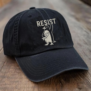 May include: Black baseball cap with the word "RESIST" embroidered above a penguin graphic holding a fish. The penguin and text are in a light color, contrasting with the dark cap. The cap has a curved brim and a fabric strap.
