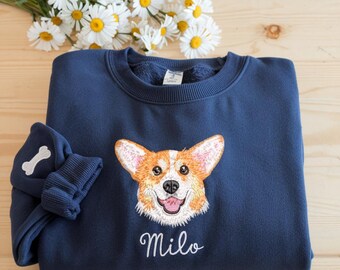 May include: Navy blue sweatshirt featuring an embroidered corgi dog face with the name "Milo" in white script. The sweatshirt has a matching cuff with a bone design. A bouquet of white and yellow daisies rests in the background.