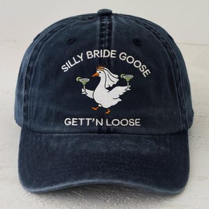 May include: Navy blue baseball cap with the embroidered text "SILLY BRIDE GOOSE GETT'N LOOSE". The cap features a white goose wearing a veil and holding two margarita glasses. The cap has a curved brim and a button on top.