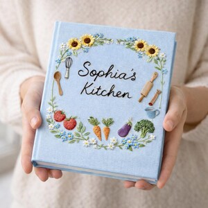May include: A light blue cookbook titled "Sophia's Kitchen" with embroidered floral and kitchen utensil details. The cover features sunflowers, daisies, strawberries, carrots, an eggplant, broccoli, a whisk, a spoon, a rolling pin, and a teacup.