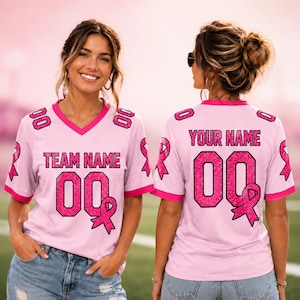 May include: A light pink V-neck jersey featuring dark pink glitter-style text that reads TEAM NAME and YOUR NAME, with the number 00 and a pink ribbon graphic on the front and back. The shirt has dark pink trim on the sleeves and neckline.