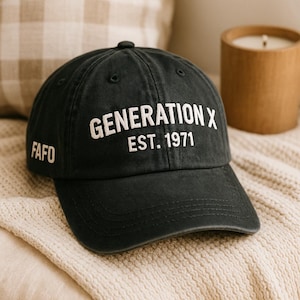 May include: Black baseball cap with the text "GENERATION X EST. 1971" in white across the front. The left side of the cap has the word "FAFO" in white. The cap is sitting on a cream-colored blanket.