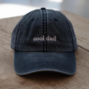 May include: A dark blue baseball cap with the embroidered text "cool dad" in a lighter shade. The cap has a curved brim and a classic six-panel design, suitable for casual wear.
