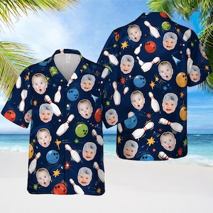 May include: A short-sleeved button-down shirt in dark blue featuring an all-over print of bowling pins, bowling balls, yellow stars, and repeated photographic portraits of a baby wearing a light blue headband against a dark blue background.