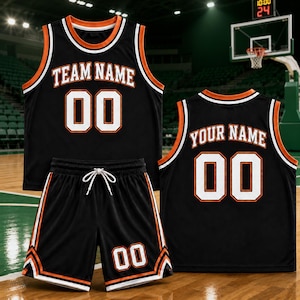 Pode incluir: Um conjunto de basquete preto composto por uma regata e shorts com detalhes em laranja e branco. A regata exibe o texto TEAM NAME e o número 00 em branco com contornos laranja. O short apresenta o número 00 e um cordão de ajuste.