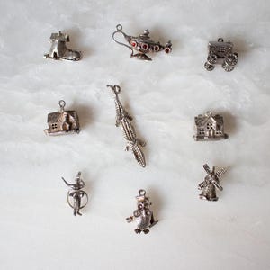 May include: A collection of nine silver charm pendants, including a house, a boot, a genie lamp, a wagon, a crocodile, a windmill, a house, a bird, and a house. The charms are all detailed and intricate.