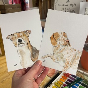 Puede incluir: Dos pinturas de acuarela de perros. Una presenta un perro marr&oacute;n y blanco, la otra un perro con manchas marrones. Ambas est&aacute;n sobre papel blanco. Las pinturas se sostienen en una mano.