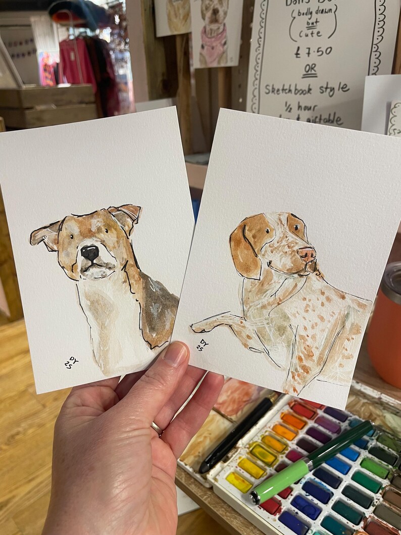 Puede incluir: Dos pinturas de acuarela de perros. Una presenta un perro marr&oacute;n y blanco, la otra un perro con manchas marrones. Ambas est&aacute;n sobre papel blanco. Las pinturas se sostienen en una mano.