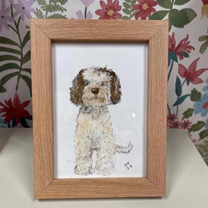 Puede incluir: Una pintura de acuarela enmarcada de un perro con pelaje marr&oacute;n y blanco. El perro est&aacute; sentado y tiene una expresi&oacute;n amigable. El marco es de madera marr&oacute;n claro. Las iniciales del artista y el a&ntilde;o, "DY 23", est&aacute;n en la esquina inferior derecha.