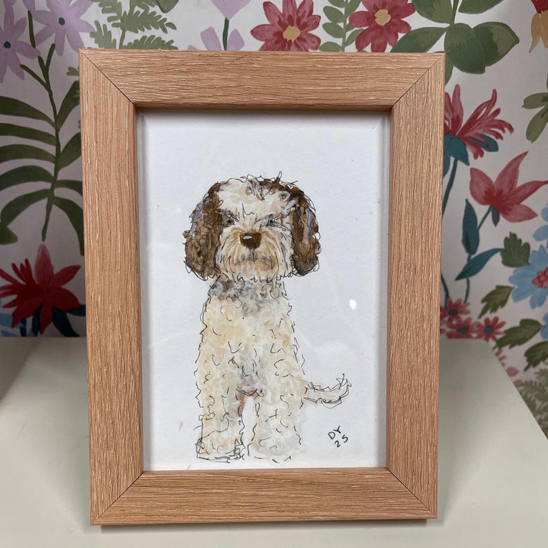 Puede incluir: Una pintura de acuarela enmarcada de un perro con pelaje marr&oacute;n y blanco. El perro est&aacute; sentado y tiene una expresi&oacute;n amigable. El marco es de madera marr&oacute;n claro. Las iniciales del artista y el a&ntilde;o, "DY 23", est&aacute;n en la esquina inferior derecha.