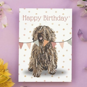 May include: A birthday card with a Puli dog illustration. The card reads "Happy Birthday" in a light brown font. The background is white with gold polka dots. A string of colourful pennants is above the dog, adding a festive touch.