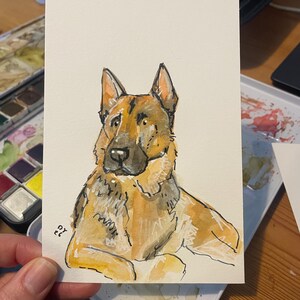 Puede incluir: Pintura a la acuarela de un perro pastor alem&aacute;n. El perro est&aacute; representado en tonos marrones, beige y negro, con un contorno detallado. La obra de arte est&aacute; sobre una tarjeta rectangular blanca, con las iniciales del artista en la esquina inferior izquierda.