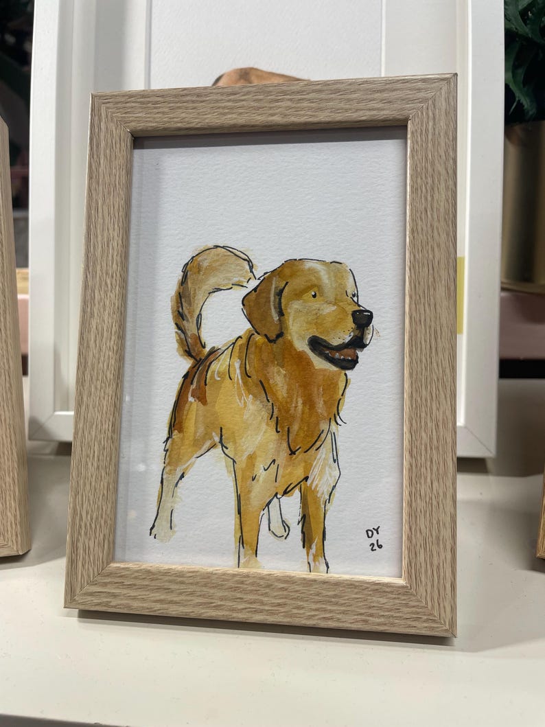 Puede incluir: Pintura a la acuarela de un perro golden retriever en un marco de madera marr&oacute;n claro. El perro est&aacute; representado en tonos dorados y marrones, con una cola esponjosa y una expresi&oacute;n feliz. La obra est&aacute; firmada "DY 26" en la esquina inferior derecha.