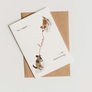 Puede incluir: Tarjeta de felicitación con sobre marrón. La tarjeta presenta dos perros ilustrados, uno marrón y blanco, el otro marrón, conectados por una cuerda roja que deletrea "love". El texto en la tarjeta dice "You tugged... my heartstrings."