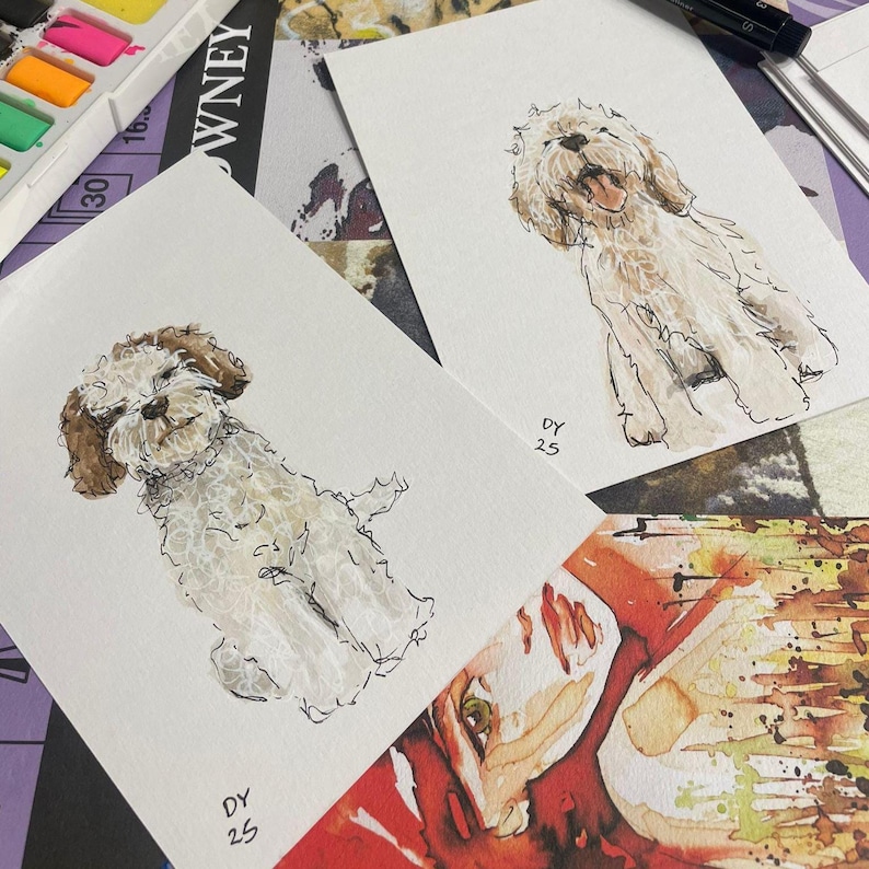 Puede incluir: Dos pinturas de acuarela de perros. Uno es marr&oacute;n claro y blanco, el otro es marr&oacute;n y blanco. Ambos est&aacute;n sentados. Las pinturas est&aacute;n sobre papel blanco con las iniciales "DY" y el n&uacute;mero "25" en la esquina. Acuarelas y un bol&iacute;grafo en el fondo.