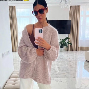 Könnte beinhalten: Eine Frau trägt einen hellrosa übergroßen Strickcardigan, ein braunes Oberteil, eine weiße Hose und beige Schuhe. Sie trägt eine rote Sonnenbrille und hält ein rosa Telefon. Der Text "Olena Riabchun" und eine ukrainische Flagge sind enthalten.