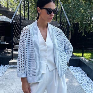 May include: A light blue, open-weave cardigan with long sleeves. The cardigan is worn over a white vest and paired with white trousers. The outfit is completed with black sunglasses. The background features a staircase and greenery.