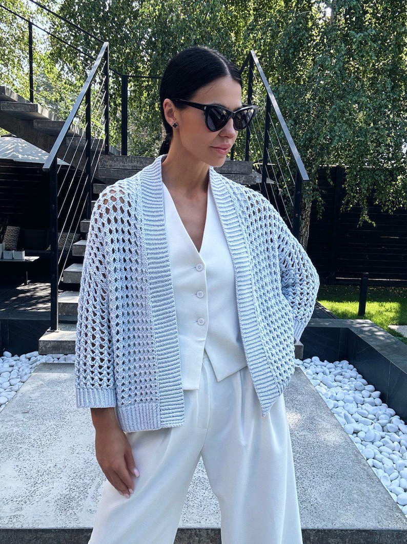 May include: A light blue, open-weave cardigan with long sleeves. The cardigan is worn over a white vest and paired with white trousers. The outfit is completed with black sunglasses. The background features a staircase and greenery.