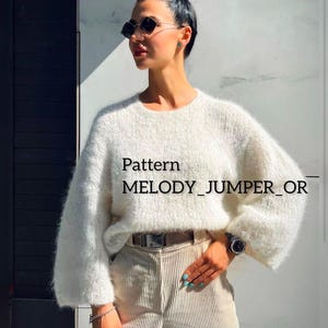 May include: A white fluffy jumper with a round neckline and long sleeves. The jumper has the text "Pattern MELODY_JUMPER_OR" printed on it. The model is wearing the jumper with a brown belt and beige corduroy trousers.