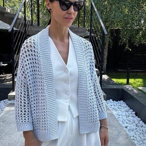 May include: A light blue open-weave cardigan with three-quarter sleeves. The cardigan is worn over a white vest and paired with white pants. The outfit is accessorized with black sunglasses and a bracelet. The background includes a staircase and greenery.