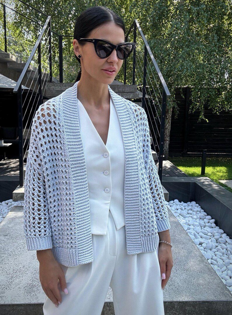 May include: A light blue open-weave cardigan with three-quarter sleeves. The cardigan is worn over a white vest and paired with white pants. The outfit is accessorized with black sunglasses and a bracelet. The background includes a staircase and greenery.