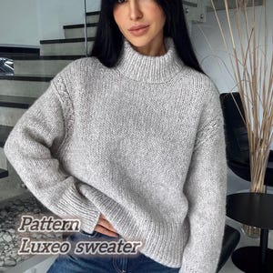 May include: A light gray, turtleneck sweater with the text "Pattern Luxeo sweater" in a casual font. The sweater has a ribbed collar and cuffs, and a textured knit design. The model is wearing blue jeans.