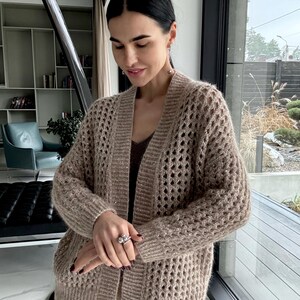 May include: A tan open-front cardigan with a textured, open-knit design. The long-sleeved sweater has a V-neck and is worn over a brown top and tan pants. The cardigan is made of a soft, possibly wool, material.