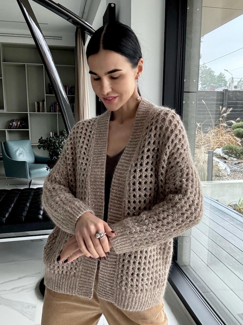May include: A tan open-front cardigan with a textured, open-knit design. The long-sleeved sweater has a V-neck and is worn over a brown top and tan pants. The cardigan is made of a soft, possibly wool, material.