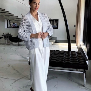 May include: A light blue open-weave cardigan over a white top and wide-leg pants. The outfit is paired with sandals. The setting is a modern interior with marble flooring and a black bench.