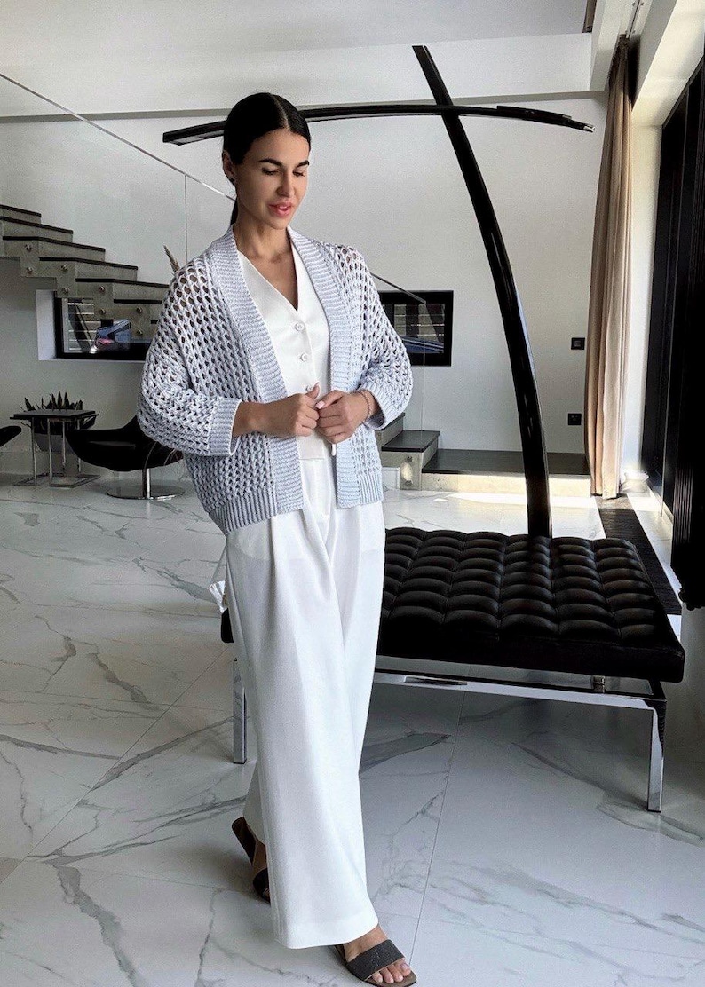 May include: A light blue open-weave cardigan over a white top and wide-leg pants. The outfit is paired with sandals. The setting is a modern interior with marble flooring and a black bench.