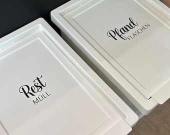 May include: Two white rectangular recycling bins with lids. One bin is labelled "Rest Müll" in a black script font, and the other is labelled "Pfand FLASCHEN". The bins are on a light brown wooden floor.