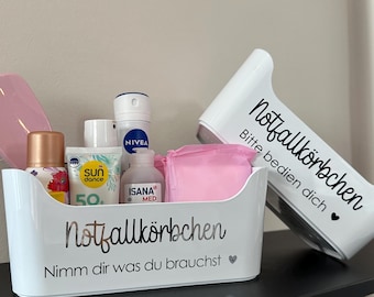 May include: A white container with curved edges, labelled "Notfallkörbchen" and "Nimm dir was du brauchst", filled with toiletries. A matching white, angled container with the text "Notfallkörbchen Bitte bedien dich" is next to it.